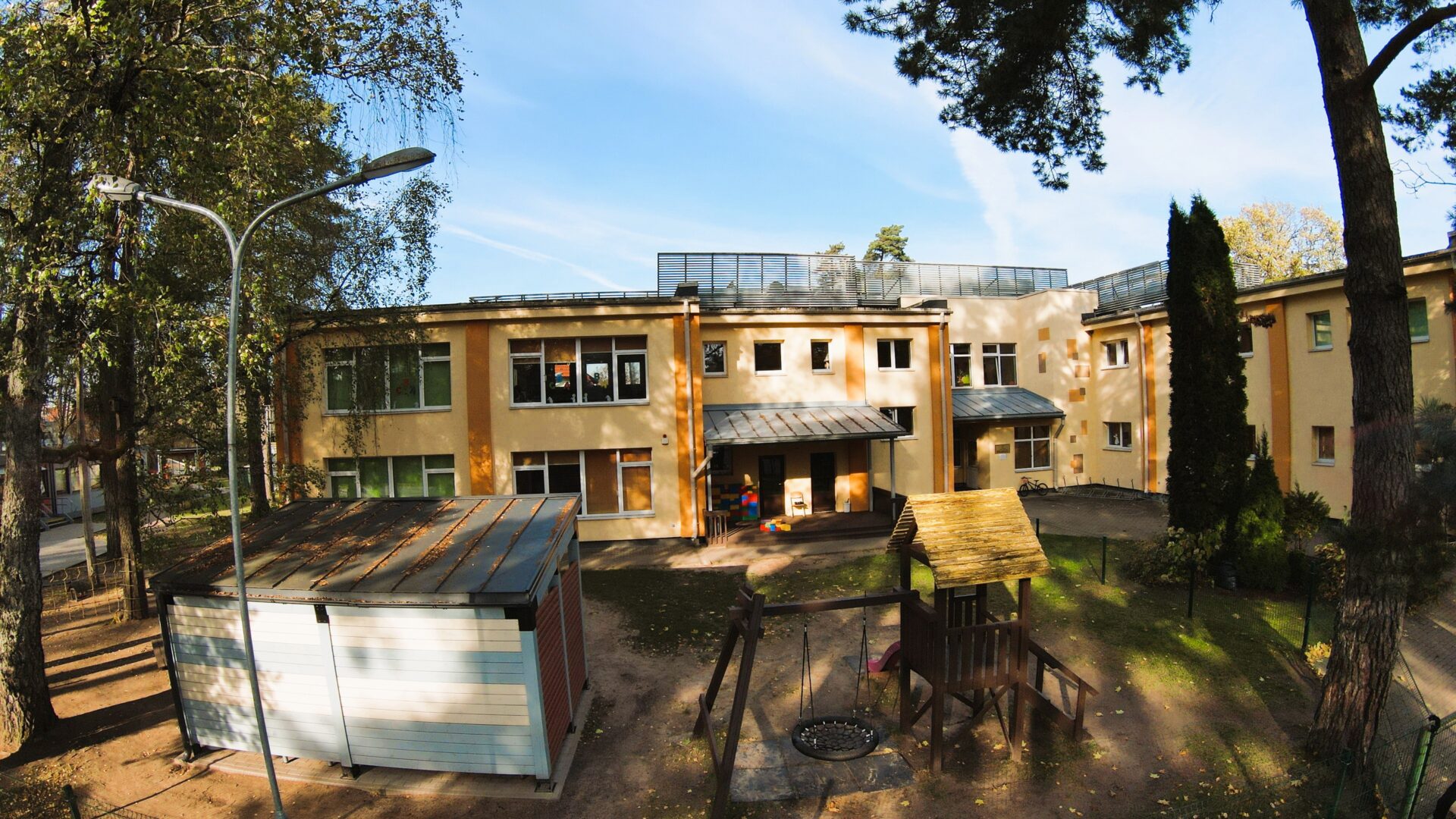 Dzeltena ēka, zemes seguma bērnu laukums, šūpoles, nojume.