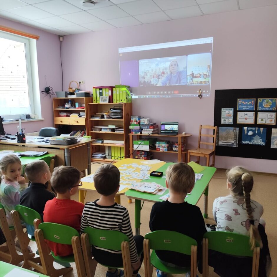 Skolotāja, bērni skatās ekrāna projekcijā uz sienas. Fonā Sprīdīšu grupas iekārtojums - plaukti, galdi, spēles.