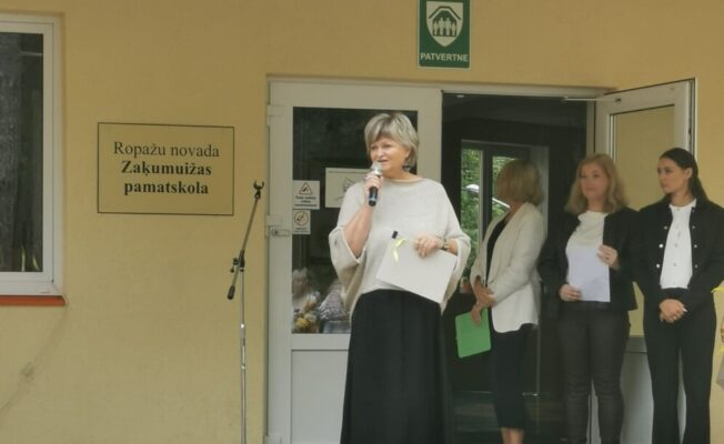 direktore runā mikrofonā, 3 pieaugušie stāvfonā. pie dzeltenas skolas ēkas ar uzrakstu Zaķumuižas pamatskola.