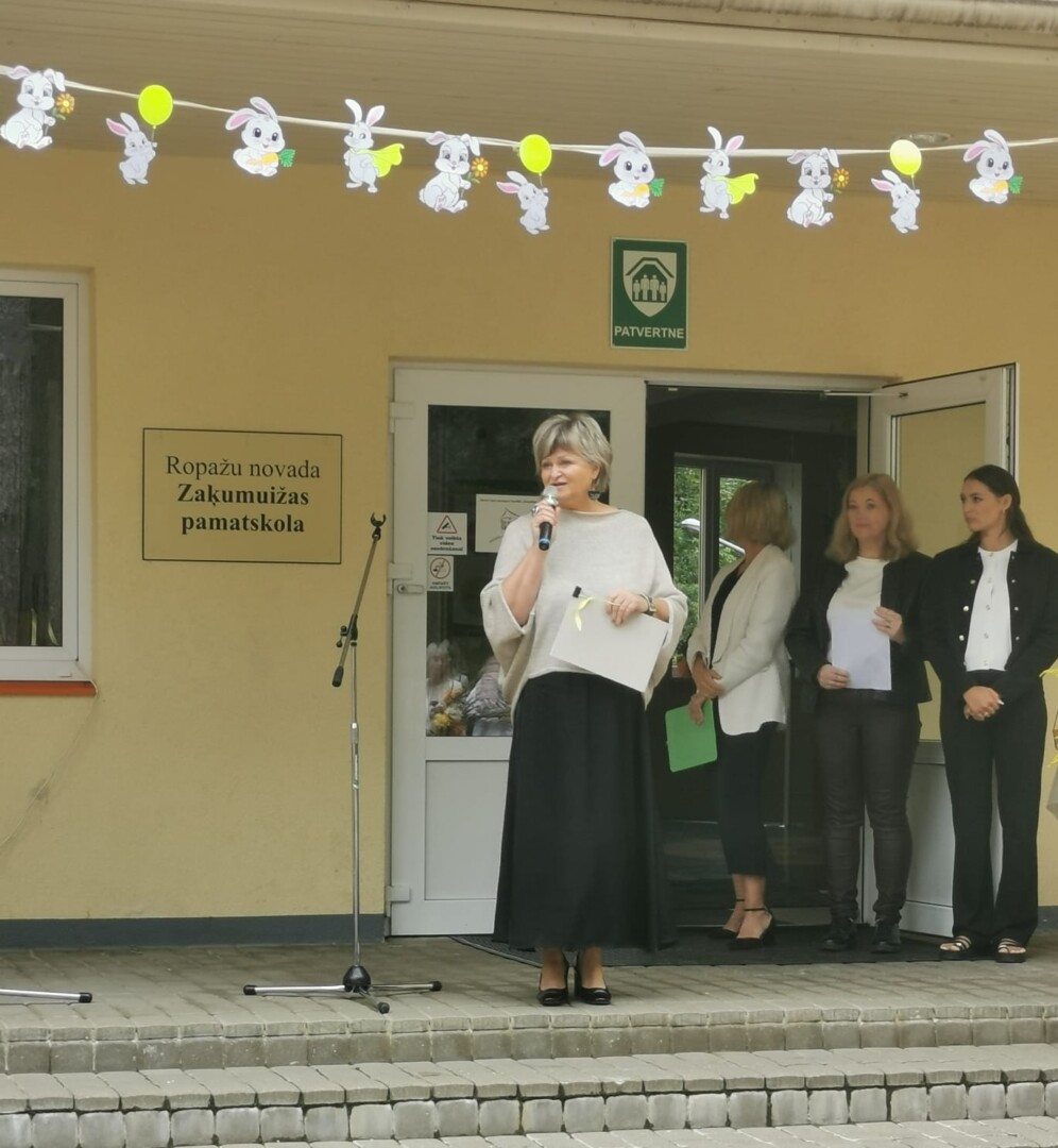 direktore runā mikrofonā, 3 pieaugušie stāvfonā. pie dzeltenas skolas ēkas ar uzrakstu Zaķumuižas pamatskola.