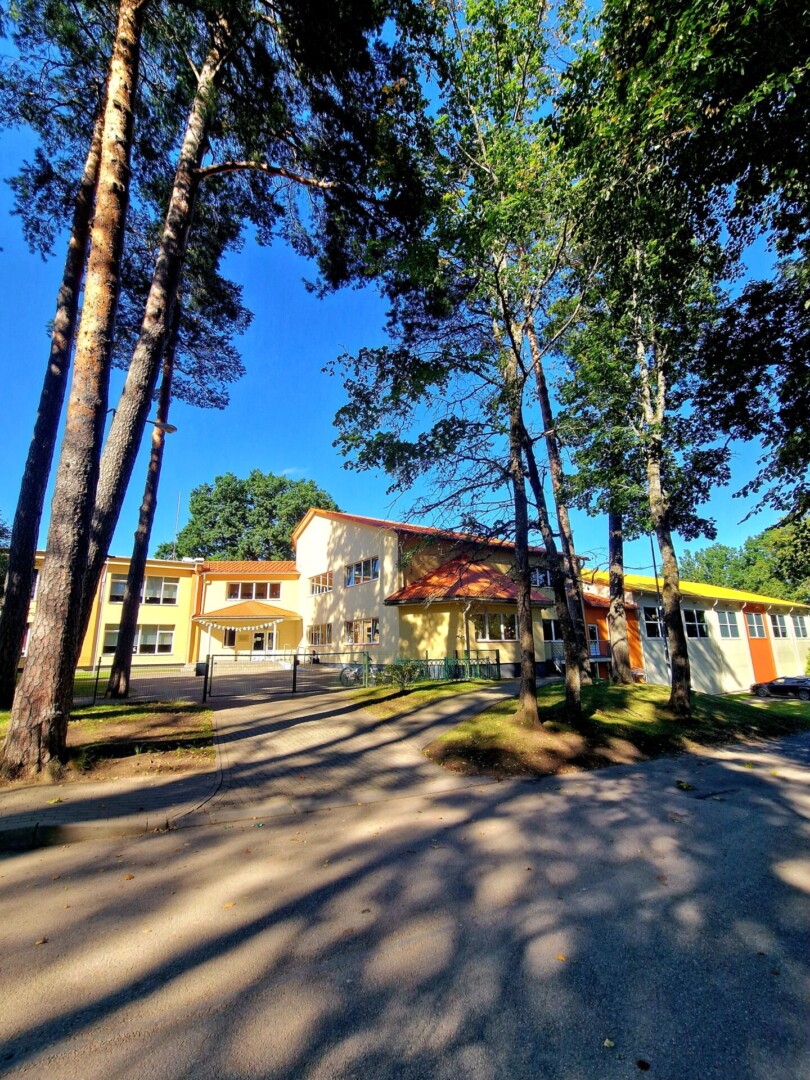 Daudzstāvu dzeltena ēka ar oranžiem akcentiem un brūni-oranžiem jumtiem. Priekšplānā asfaltēts ceļš ar garām koku ēnām. Sānos redzami augsti koki, fonā gaiši zilas debesis.
