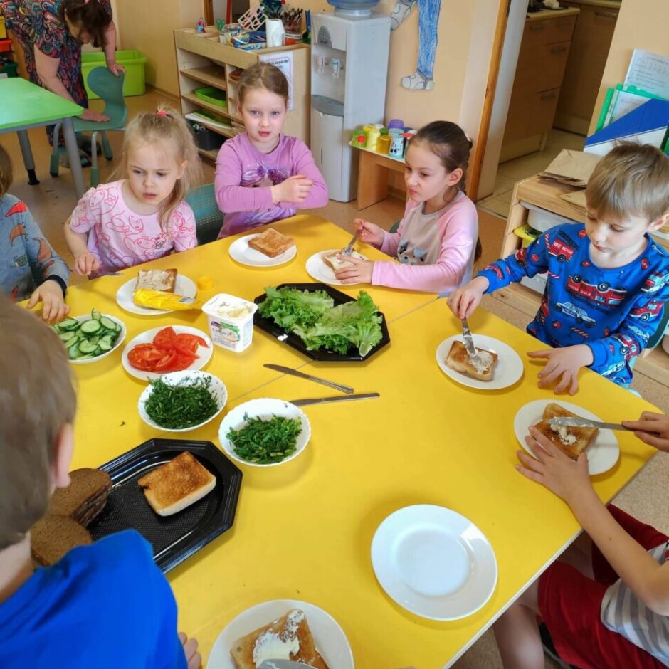 Bērni pie dzeltena galda gatavo sviestmaizes. Viņi smērē sviestu vai krēmsieru uz grauzdiņiem, uz galda ir bļodas ar sagrieztiem gurķiem, tomātiem, salātiem un dillēm. Fonā redzama skolotāja.