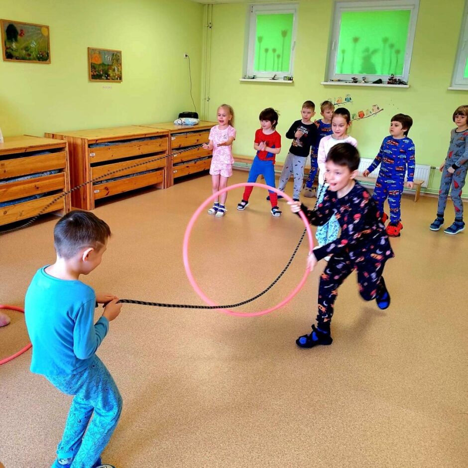 Divi zēni rotaļājas ar virvi un rozā hula hop riņķi, kamēr citi bērni viņus vēro gaišā rotaļu telpā. Viens zēns velk virvi, bet otrs skrien, turot riņķi. Telpai ir zaļas sienas un koka plaukti.