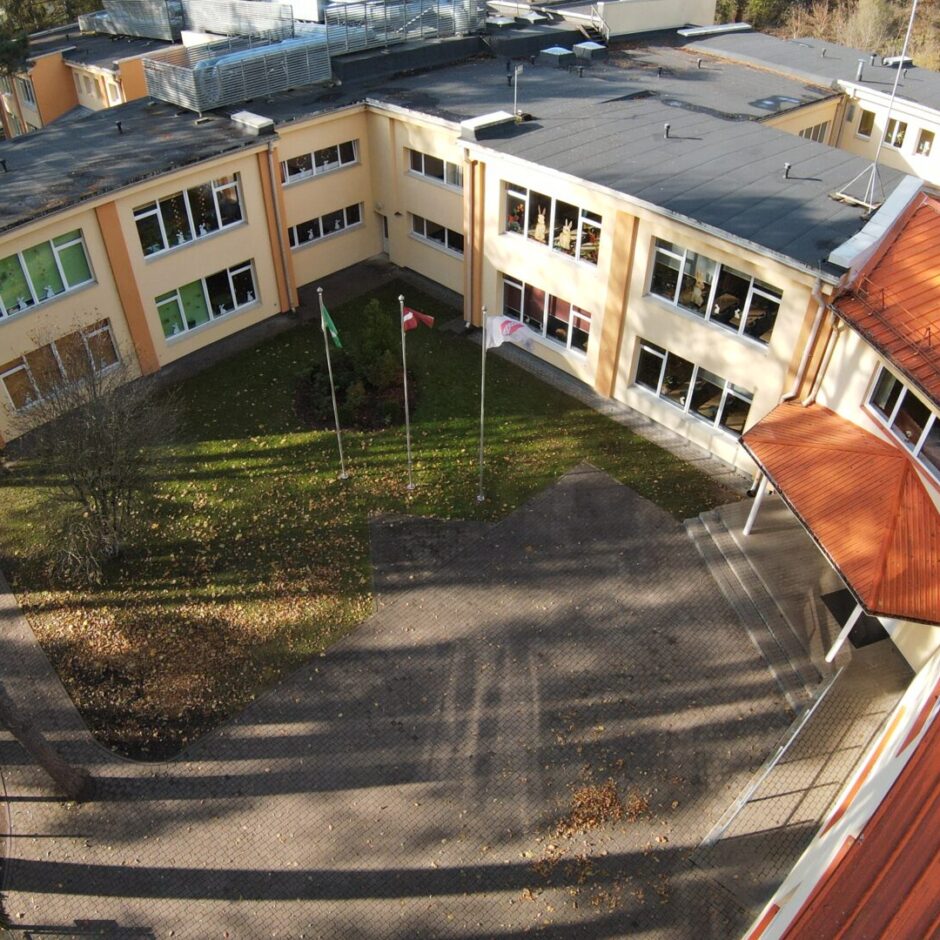 Skats no augšas uz gaišdzeltenu skolas ēku ar tumšiem un oranžīgiem jumtiem. Pagalmā redzams zāliens ar rudens lapām un bruģēta laukums, kurā mastos plīvo zaļš Ekoskolu karogs, Latvijas karogs un balts Ropažu novada karogs ar logotipu. Kreisajā pusē aug priedes.