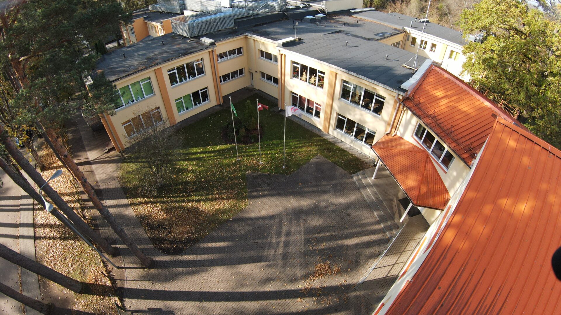 Skats no augšas uz gaišdzeltenu skolas ēku ar tumšiem un oranžīgiem jumtiem. Pagalmā redzams zāliens ar rudens lapām un bruģēta laukums, kurā mastos plīvo zaļš Ekoskolu karogs, Latvijas karogs un balts Ropažu novada karogs ar logotipu. Kreisajā pusē aug priedes.