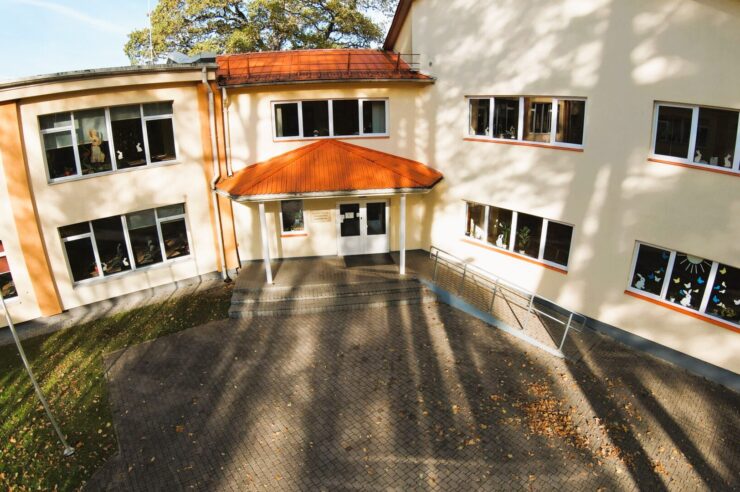 Skolas ēkas fasāde, gaiša divstāvu ēka ar oranžu jumtu virs galvenās ieejas. Logi ir rotāti ar zaķiem, tauriņiem un sauli. Pie ieejas kāpnēm ir uzbrauktuve invalīdiem. Uz zemes redzamas kritušas lapas.