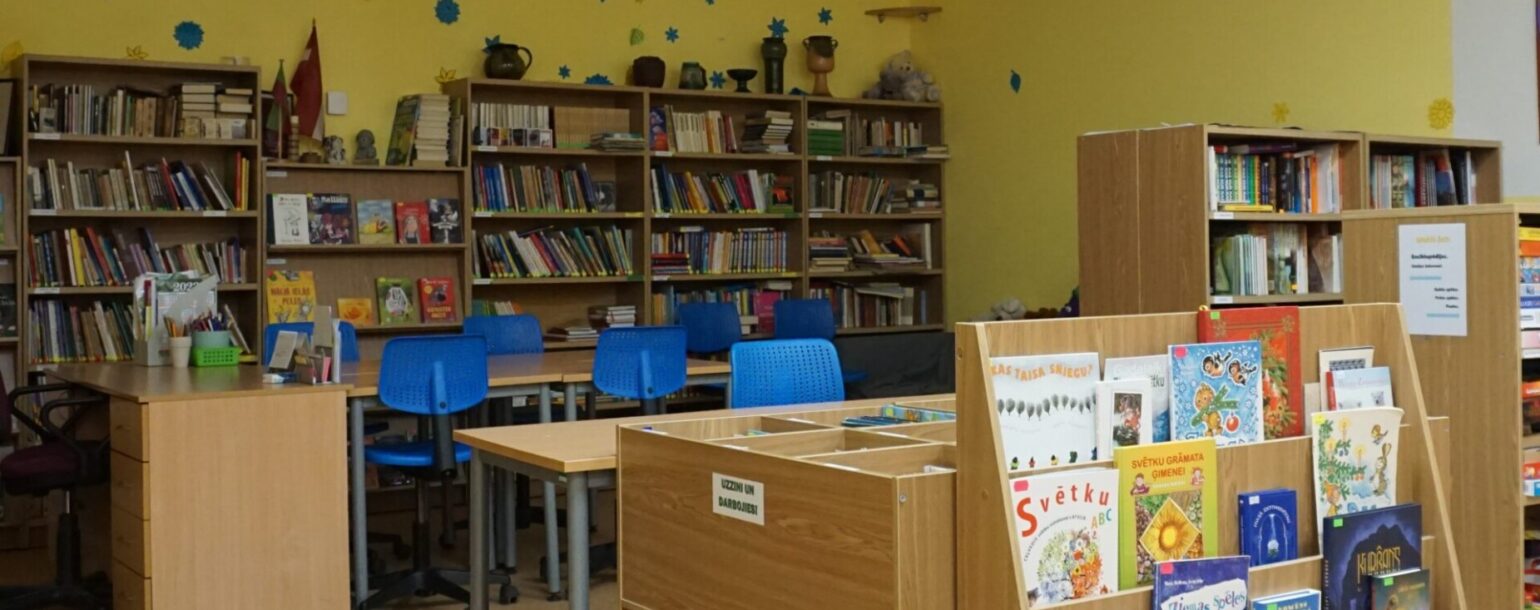 Dzeltena skolas bibliotēkas telpa ar grāmatu plauktiem, kas pilni ar grāmatām, gaišiem galdiem un ziliem krēsliem. Sienas rotā zvaigznes un sniegpārslas. Priekšplānā ir koka plaukts ar bērnu grāmatām, tostarp 