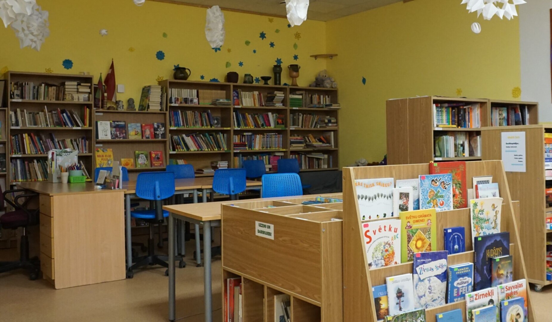 Dzeltena skolas bibliotēkas telpa ar grāmatu plauktiem, kas pilni ar grāmatām, gaišiem galdiem un ziliem krēsliem. Sienas rotā zvaigznes un sniegpārslas. Priekšplānā ir koka plaukts ar bērnu grāmatām, tostarp 