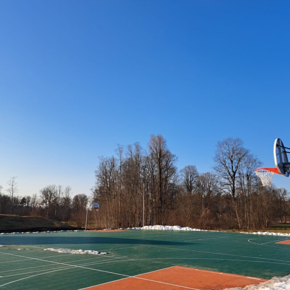 Āra basketbola laukums ar zaļu un oranžu segumu, kura malās redzami sniega pleķi. Fonā kaili koki zem dzidri zilas debess. Labajā pusē izcelts viens basketbola grozs ar oranžu tīklu, un otrs grozs redzams tālāk kreisajā pusē.