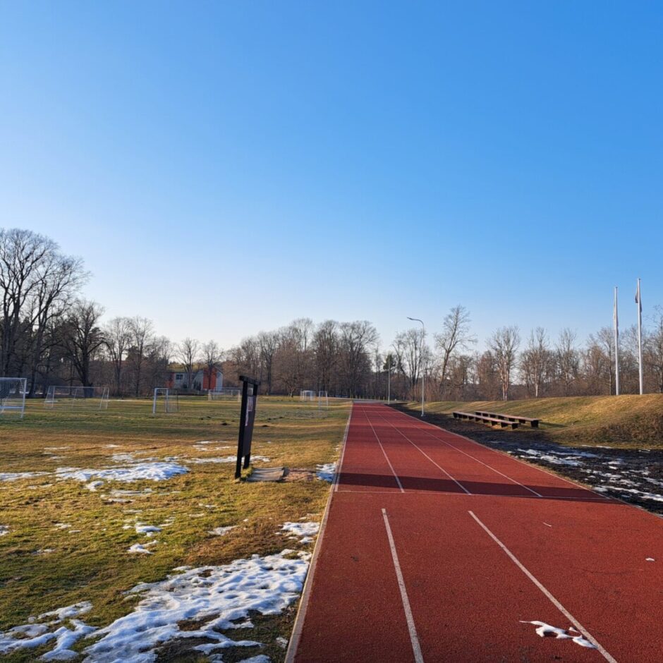 Sarkans skrejceļš ar baltām līnijām garām zaļam zālienam ar vairākiem futbola vārtiem. Zemi sedz sniega laukumi. Fonā redzami kaili koki, zilas debesis un trīs karogu masti uz neliela pakalna.