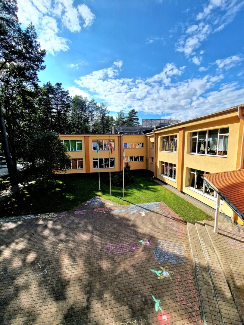 Dzeltena skolas ēka ar daudziem logiem un trim karogu mastiem. Priekšā ir bruģēts pagalms ar krāsainiem krīta zīmējumiem un koku ēnām. Apkārtnē ir zaļš zāliens un koki, bet virs ēkas – zilas debesis ar baltiem mākoņiem.