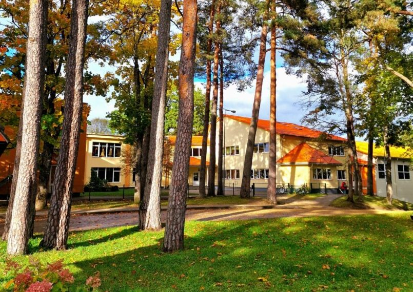 Rudens skats uz dzeltenu ēku ar oranžu jumtu, kuru ieskauj garas priedes un koki ar dzeltenām lapām, priekšplānā zaļš zāliens ar rozā ziediem.
