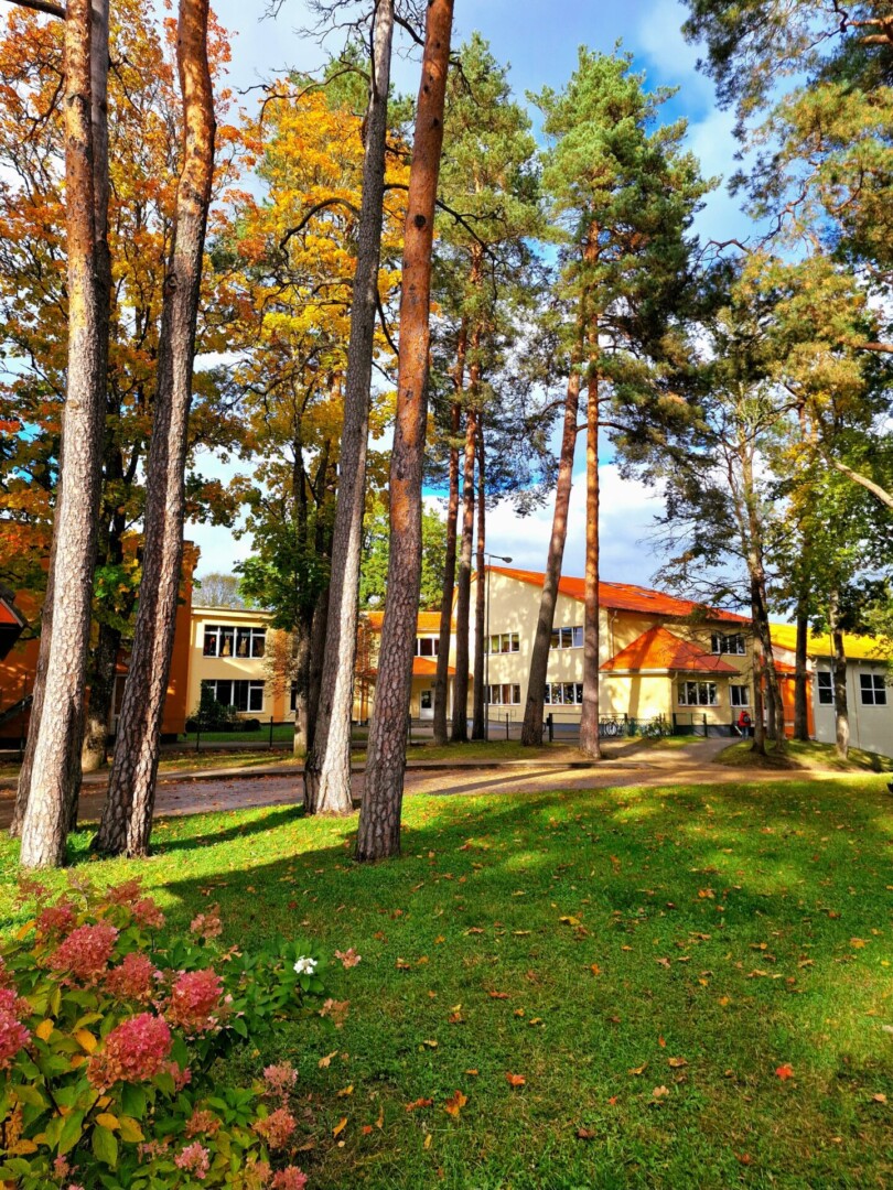 Rudens skats uz dzeltenu ēku ar oranžu jumtu, kuru ieskauj garas priedes un koki ar dzeltenām lapām, priekšplānā zaļš zāliens ar rozā ziediem.