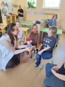 Smaidoša sieviete bērnudārza klasē nometusies ceļos, rādot rotaļu lelli zēnam, kurš sēž uz krēsla un priecīgi uz to skatās. Aiz zēna sēž divas meitenes, kas arī vēro notiekošo. Fonā redzami klases galdi un vēl viena pieaugušā.