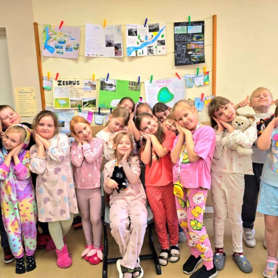 Grupa bērnu pidžamās pozē klases telpā. Daudzi bērni ir nolikuši rokas zem vaigiem, it kā gulētu. Viens zēns izkāris mēli, cits rāda īkšķus, bet meitene labajā malā aizsegusi acis.