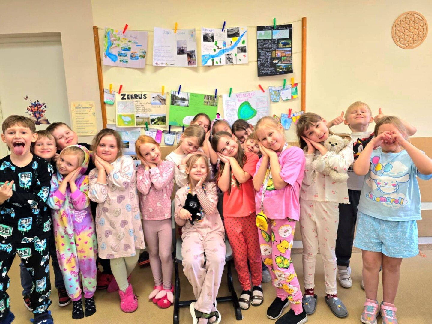 Grupa bērnu pidžamās pozē klases telpā. Daudzi bērni ir nolikuši rokas zem vaigiem, it kā gulētu. Viens zēns izkāris mēli, cits rāda īkšķus, bet meitene labajā malā aizsegusi acis.