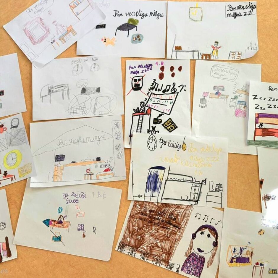 Uz koka galda izvietoti bērnu zīmējumi par veselīgu miegu, attēlojot gultas, guļošus cilvēkus, pulksteņus un miega rituālus. Vairākos zīmējumos redzams teksts 