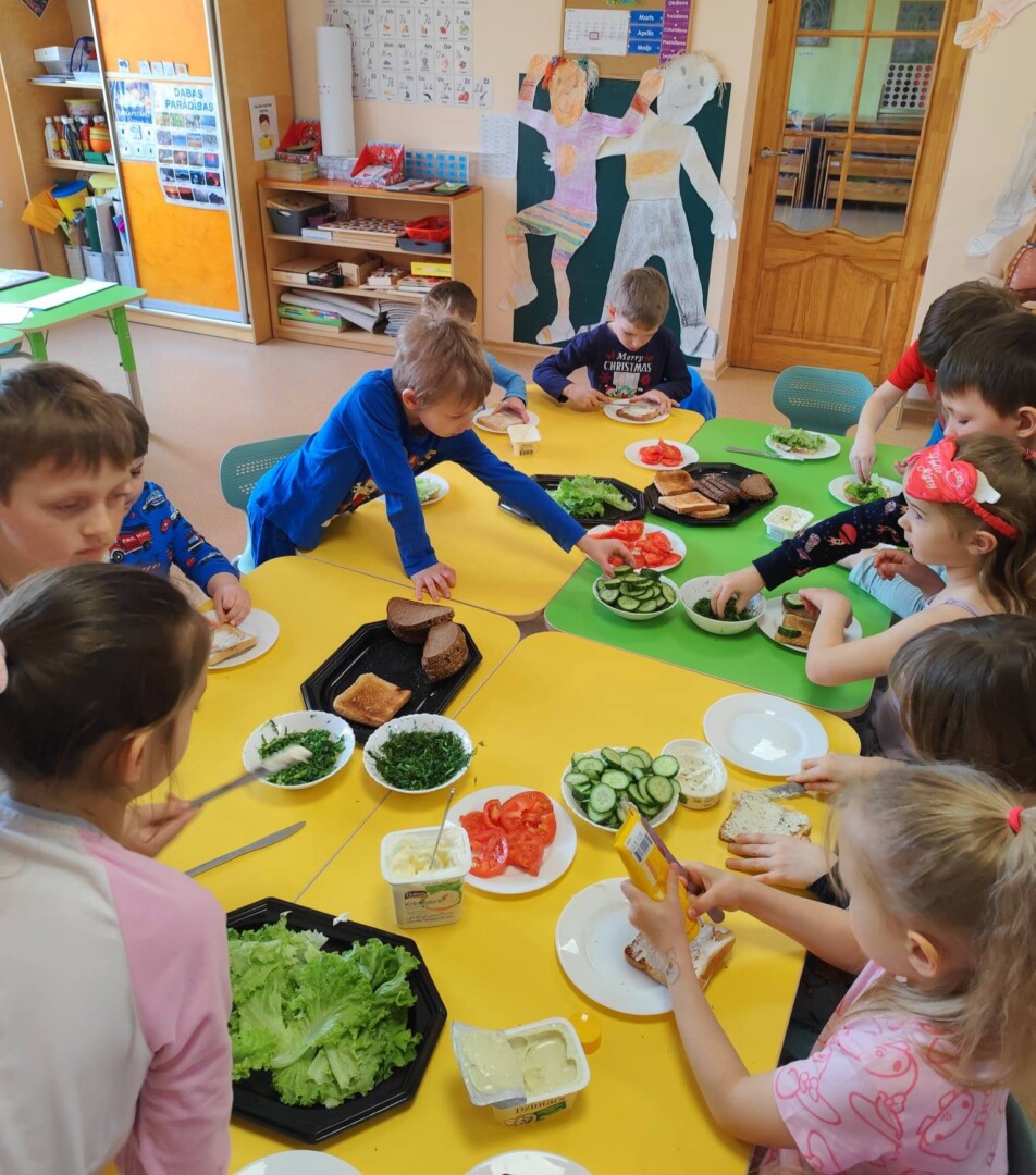 Mācību telpā bērni pie dzelteniem un zaļiem galdiem gatavo sviestmaizes, izmantojot maizi, dārzeņus un smērējumus. Viņi aktīvi liek sastāvdaļas uz maizes.