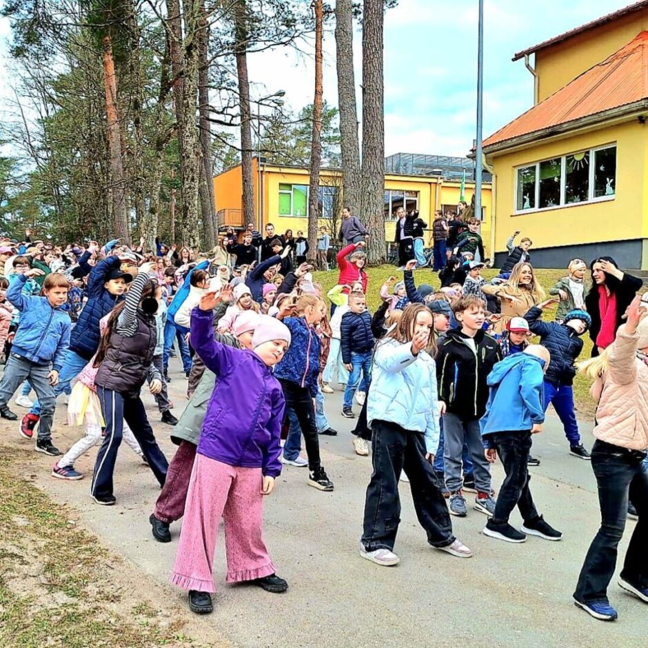 Liela bērnu grupa un daži pieaugušie ārā uz bruģēta ceļa veic sinhronus vingrinājumus ar paceltām rokām. Gar ceļu aug priedes, bet fonā redzama dzeltena skolas ēka.