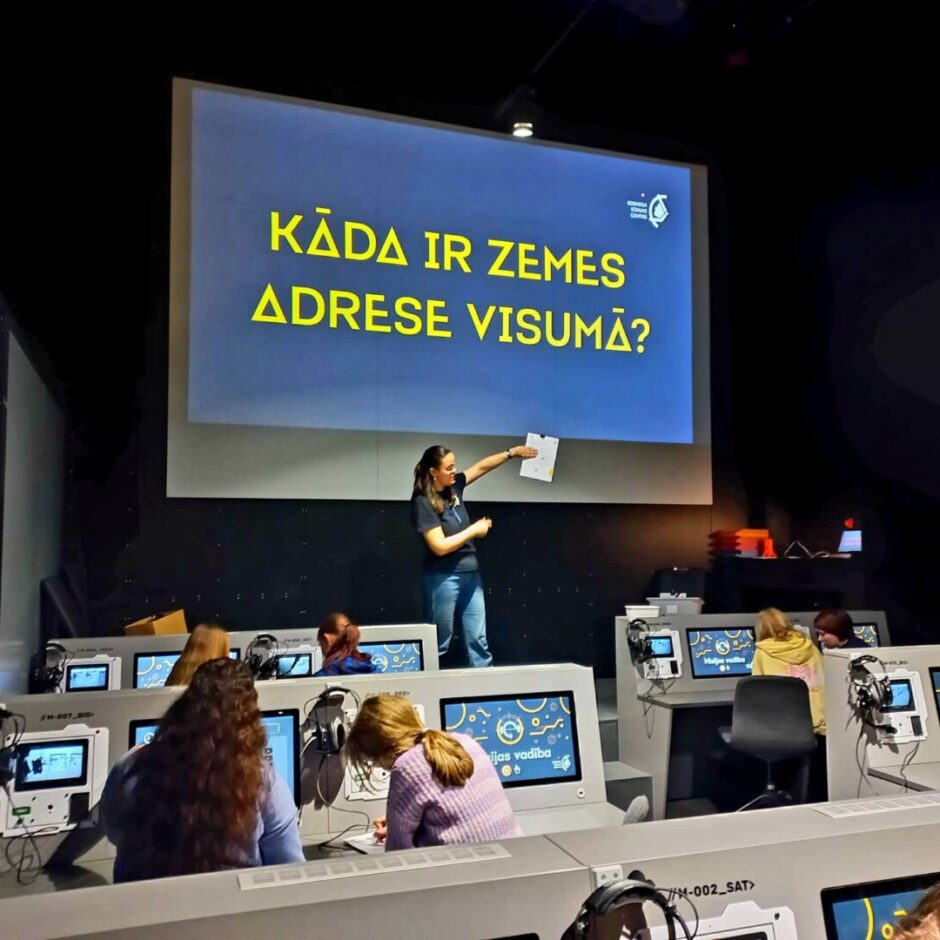 Sieviete prezentētāja stāv tumšā telpā un rāda uz lielu ekrānu, uz kura dzelteniem burtiem rakstīts 