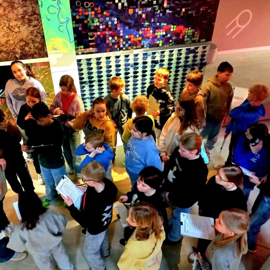 Bērnu grupa ar pieaugušajiem kosmosa zinātnes centrā. Bērni tur piezīmju lapiņas un klausās pieaugušā sievietē. Fona sienas klātas ar krāsainiem displejiem un kartēm, tostarp uzrakstiem 