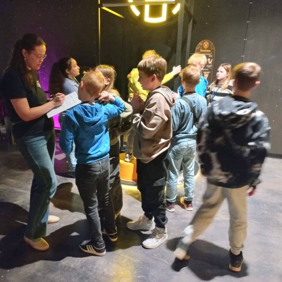Sieviete brillēs un tumšā T-kreklā raksta uz planšetes, stāvot kopā ar bērnu grupu blāvā izstāžu zālē. Puisēns zilā džemperī ar kapuci stāv viņai blakus. Fonā redzama neona dzeltena spīdoša konstrukcija un brūna zīme uz sienas ar uzrakstu 