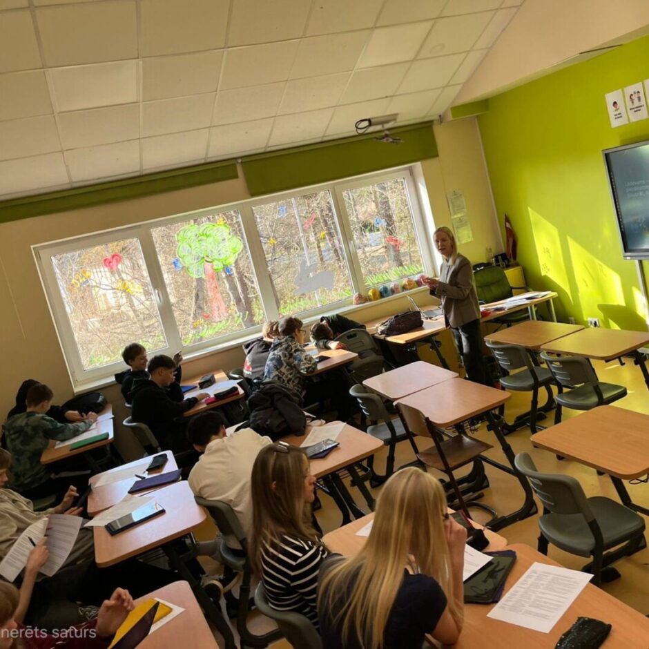Lektore stāv klases priekšpusē gaišzaļā telpā ar daudziem skolēniem, kas sēž pie individuāliem galdiem. Logi ir dekorēti ar krāsainiem zīmējumiem. Uz lielā ekrāna sienas priekšpusē redzams teksts 